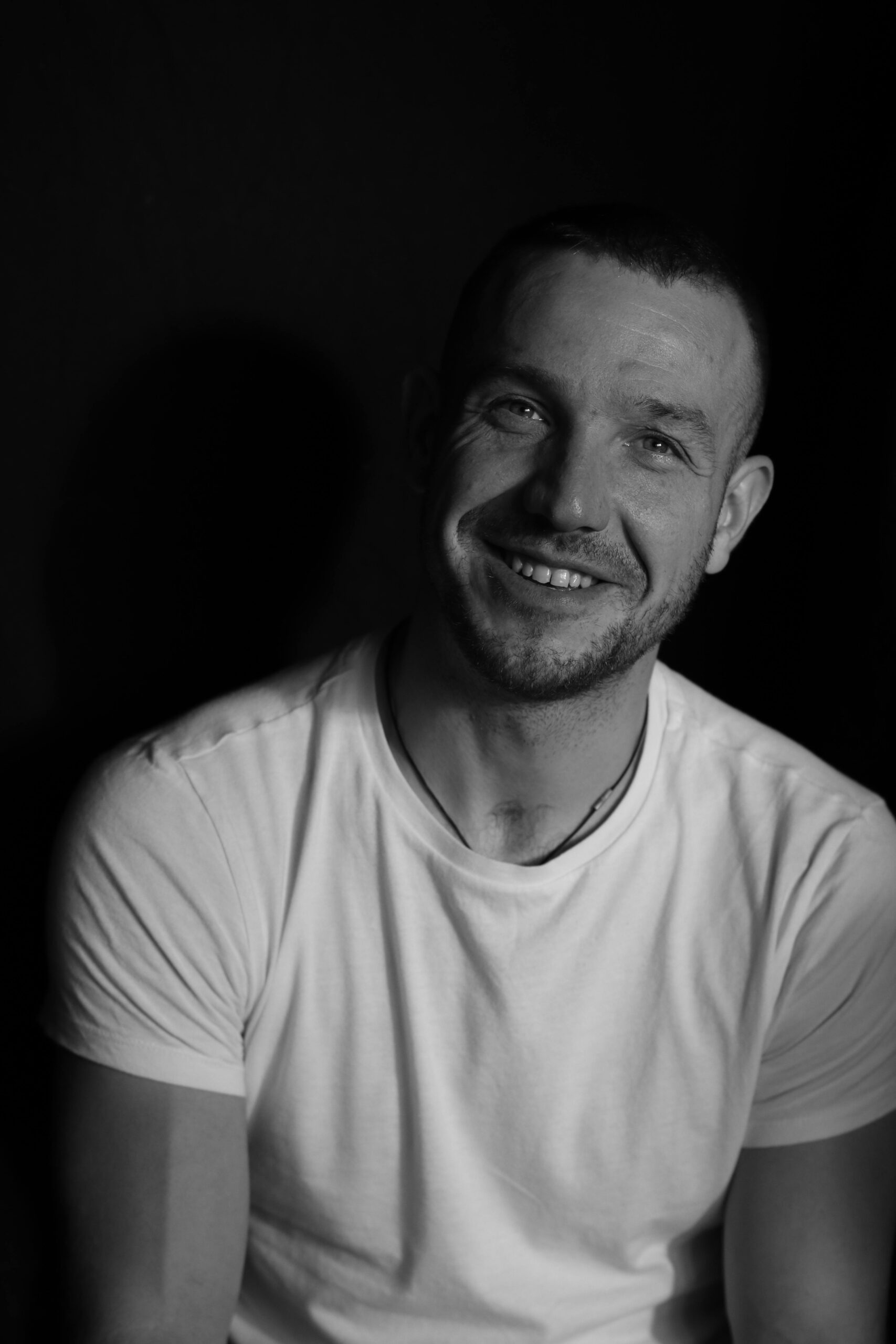 A grayscale portrait of a smiling man in a white t-shirt against a dark background.
